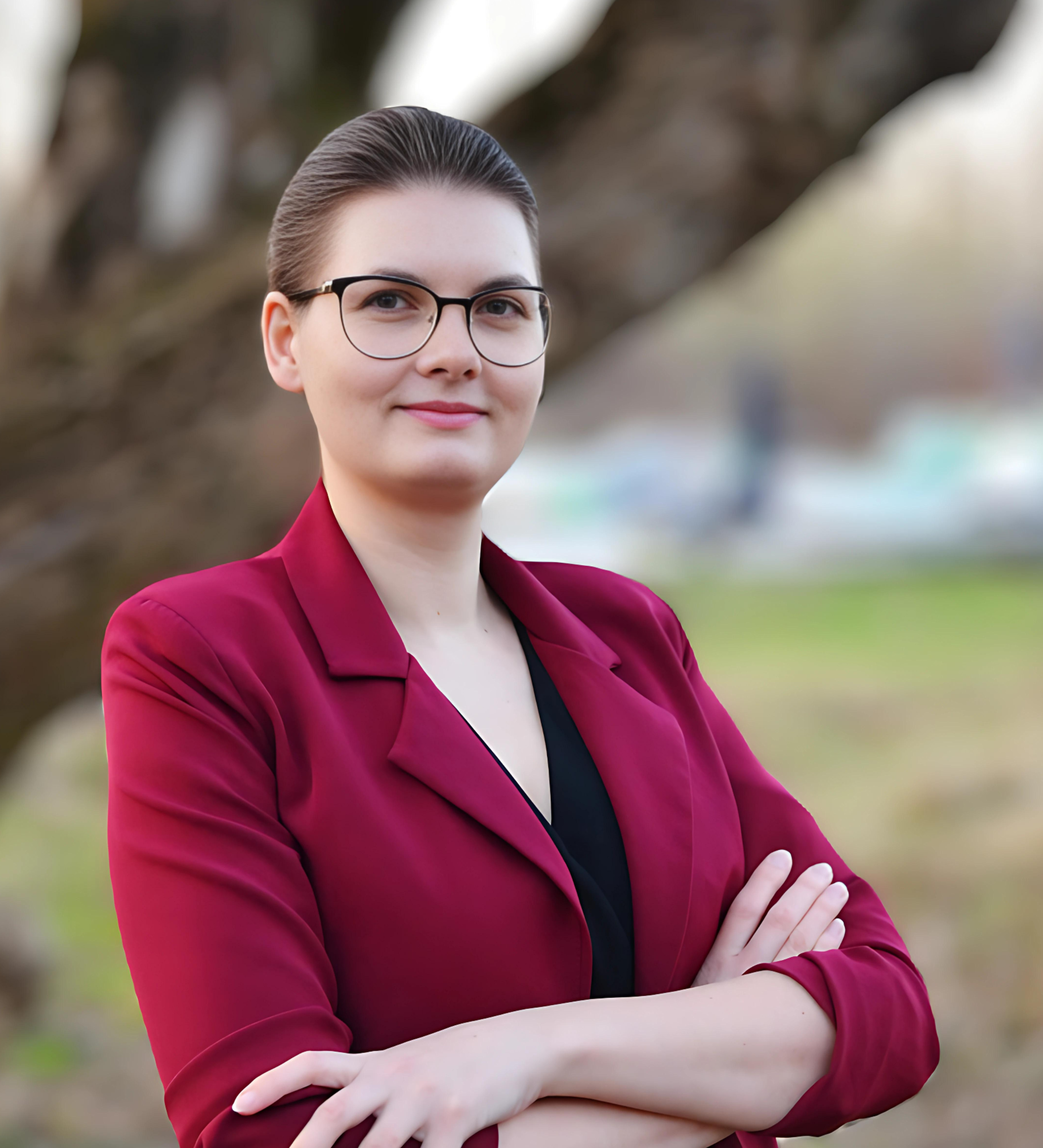 Kandydatka na radną Magdalena Tomczak na tle zieleni. Jest ubrana w czerwoną marynarkę i czarną koszulę.