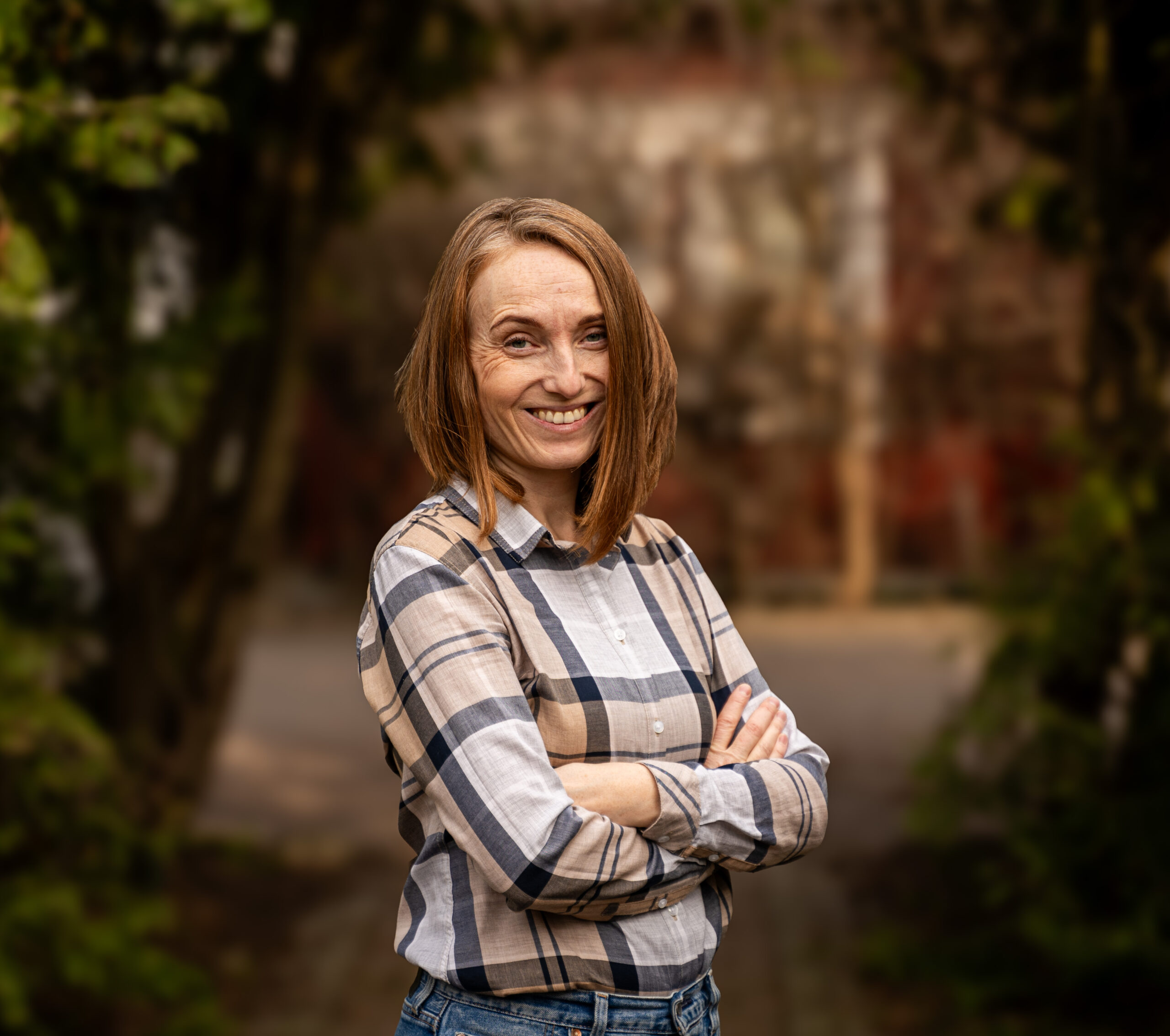 Na zdjęciu widać uśmiechniętą Paulinę Zychowicz w kraciastej koszuli. W tle widać niewyraźną obrośniętą zielenią bramę i dom.